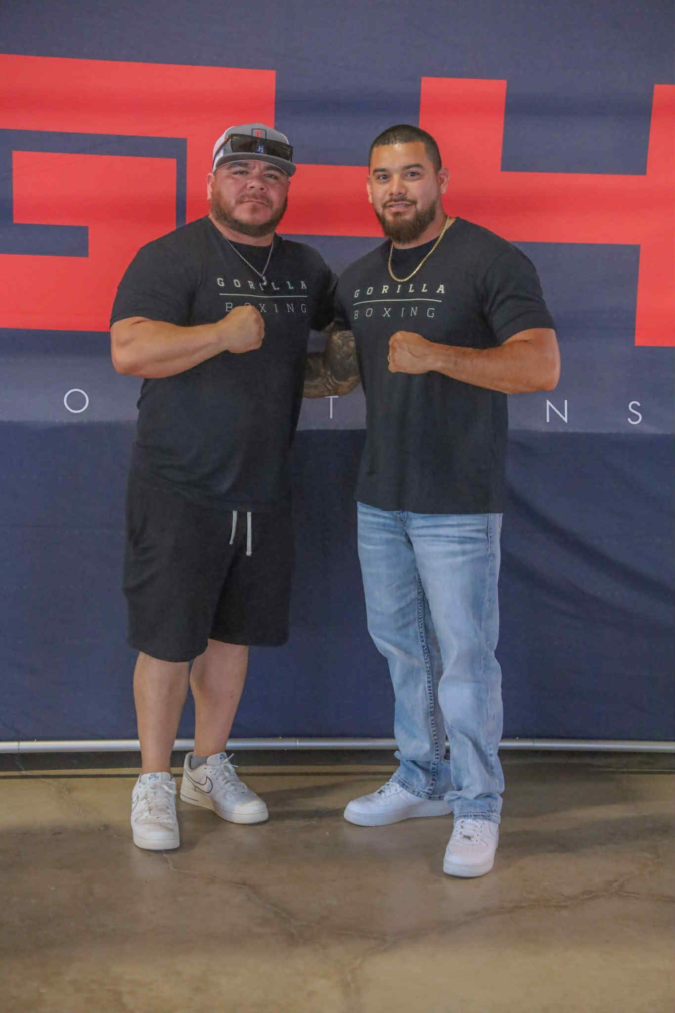 Two men in black shirts pose together in front of a dark backdrop with large red lettering.