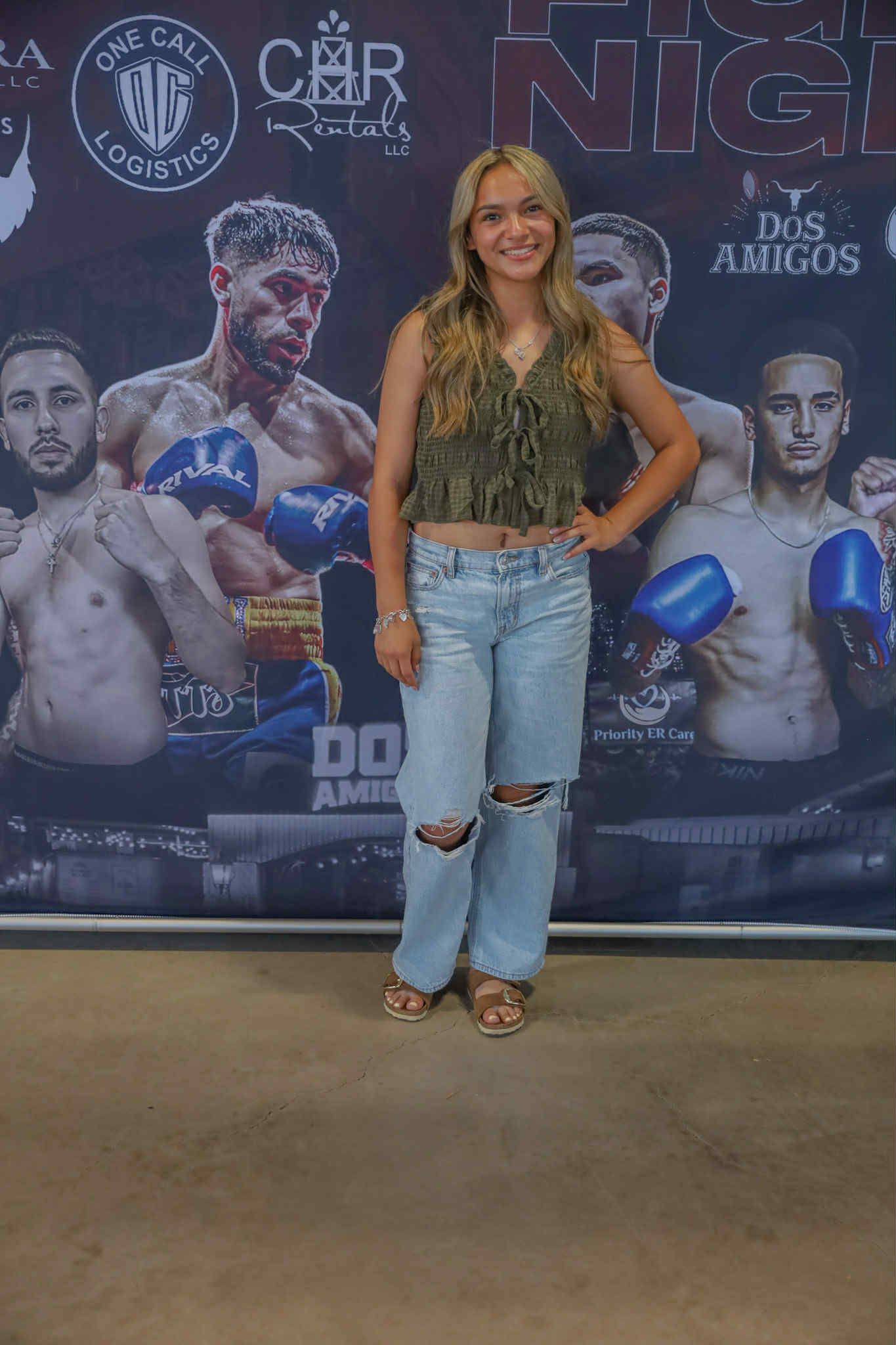 Woman standing before a boxing event backdrop, wearing a green top, light jeans, and heels.