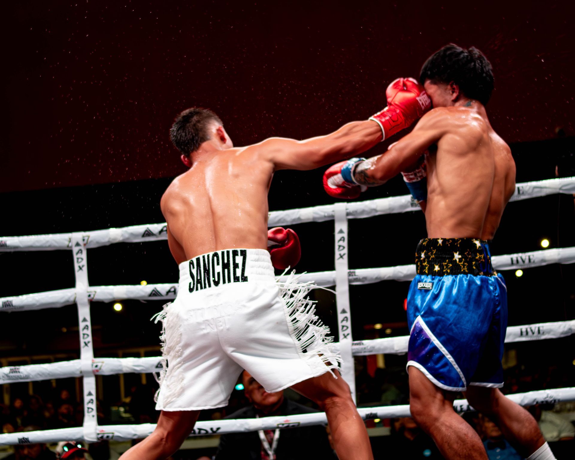Two boxers trade punches in a ring, one in white trunks and one in blue trunks.