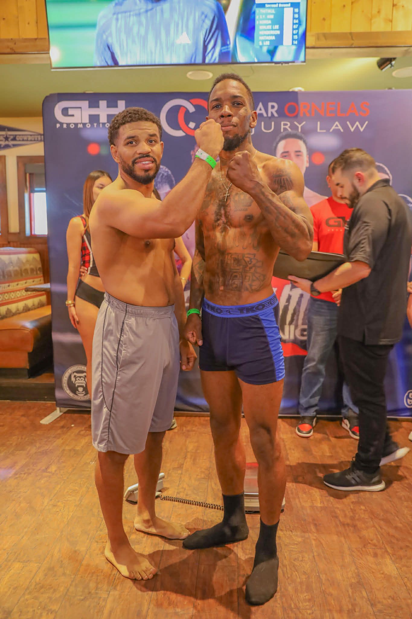 Two men pose on a stage, flexing their arms in front of a banner, with a third person in the background.