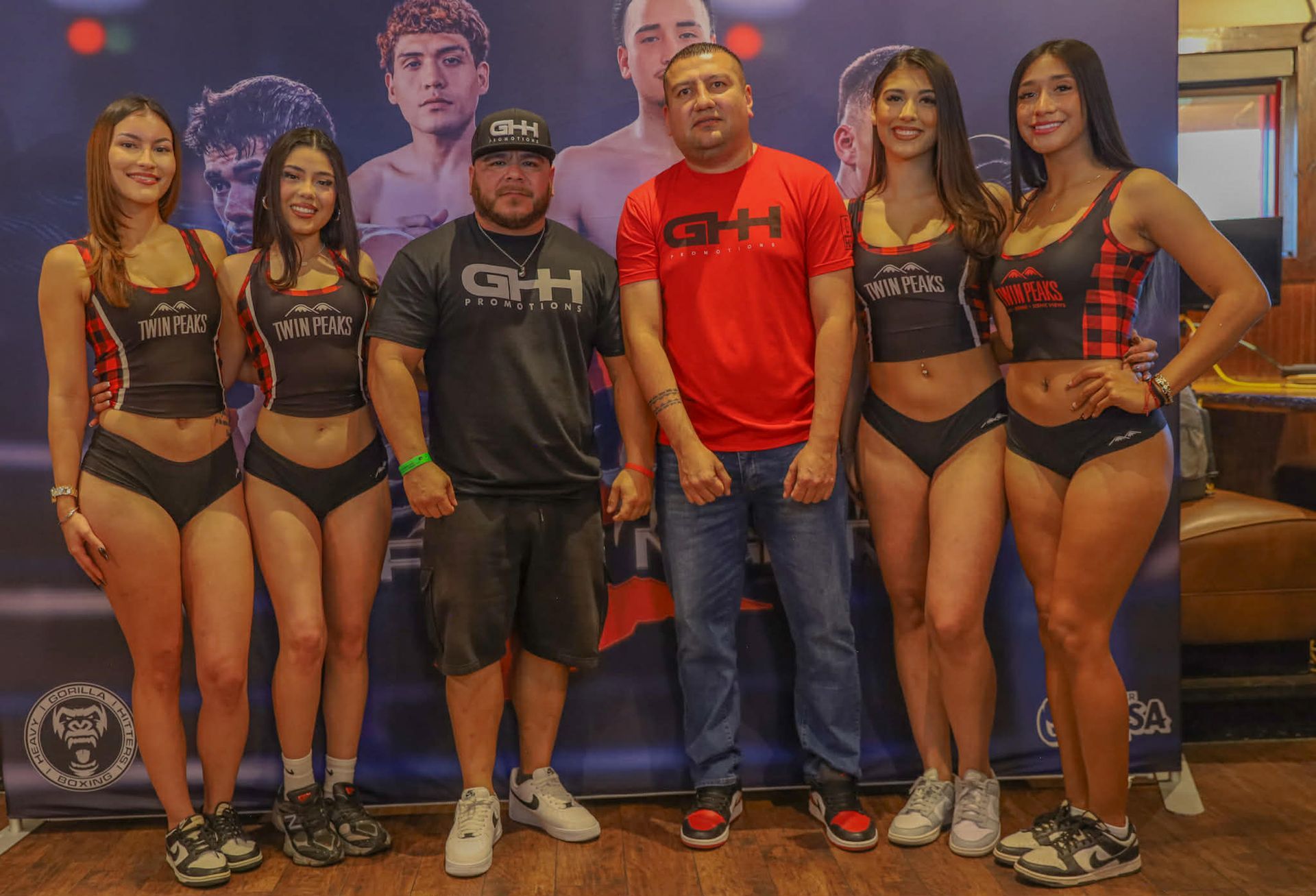 Seven people pose in front of a bodybuilding poster, wearing matching GHH shirts and competition outfits.