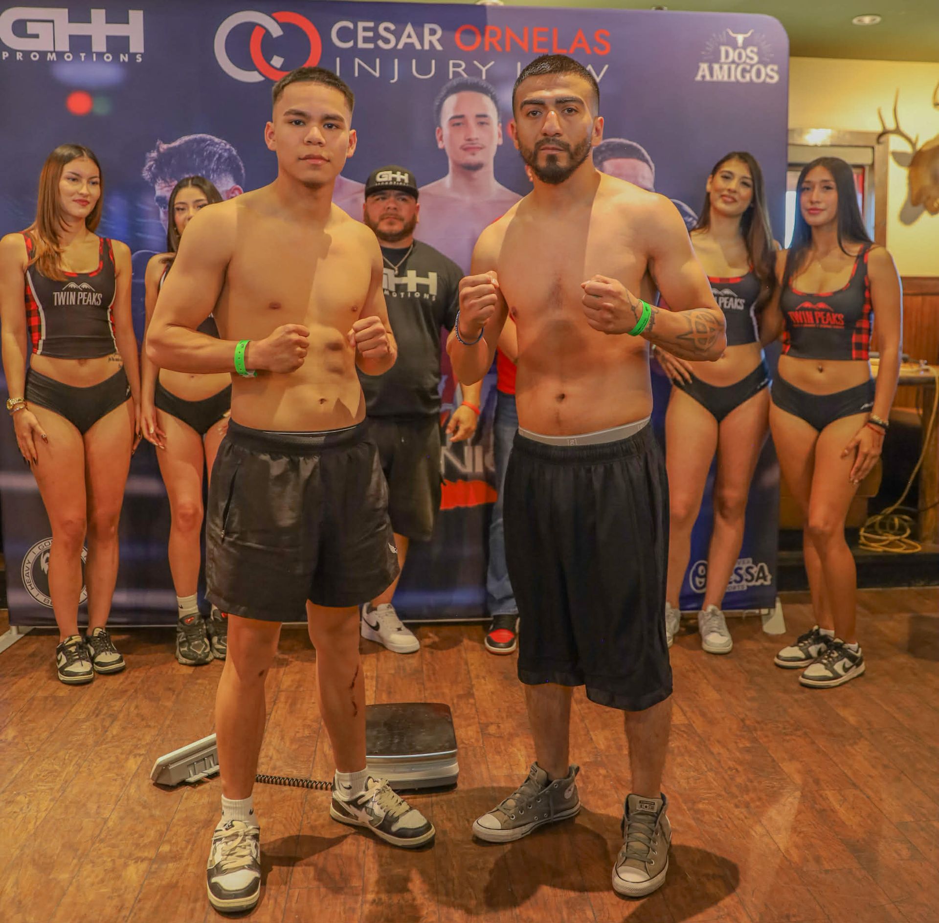 Two boxers posing at a weigh-in backdrop with ring girls and event logos behind them.