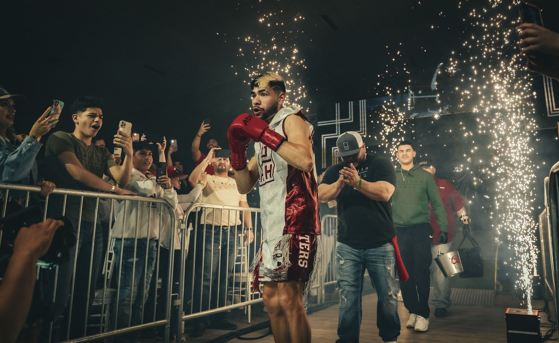Boxer in red robe celebrating in a ring as sparklers burst and a cheering crowd watches behind barriers