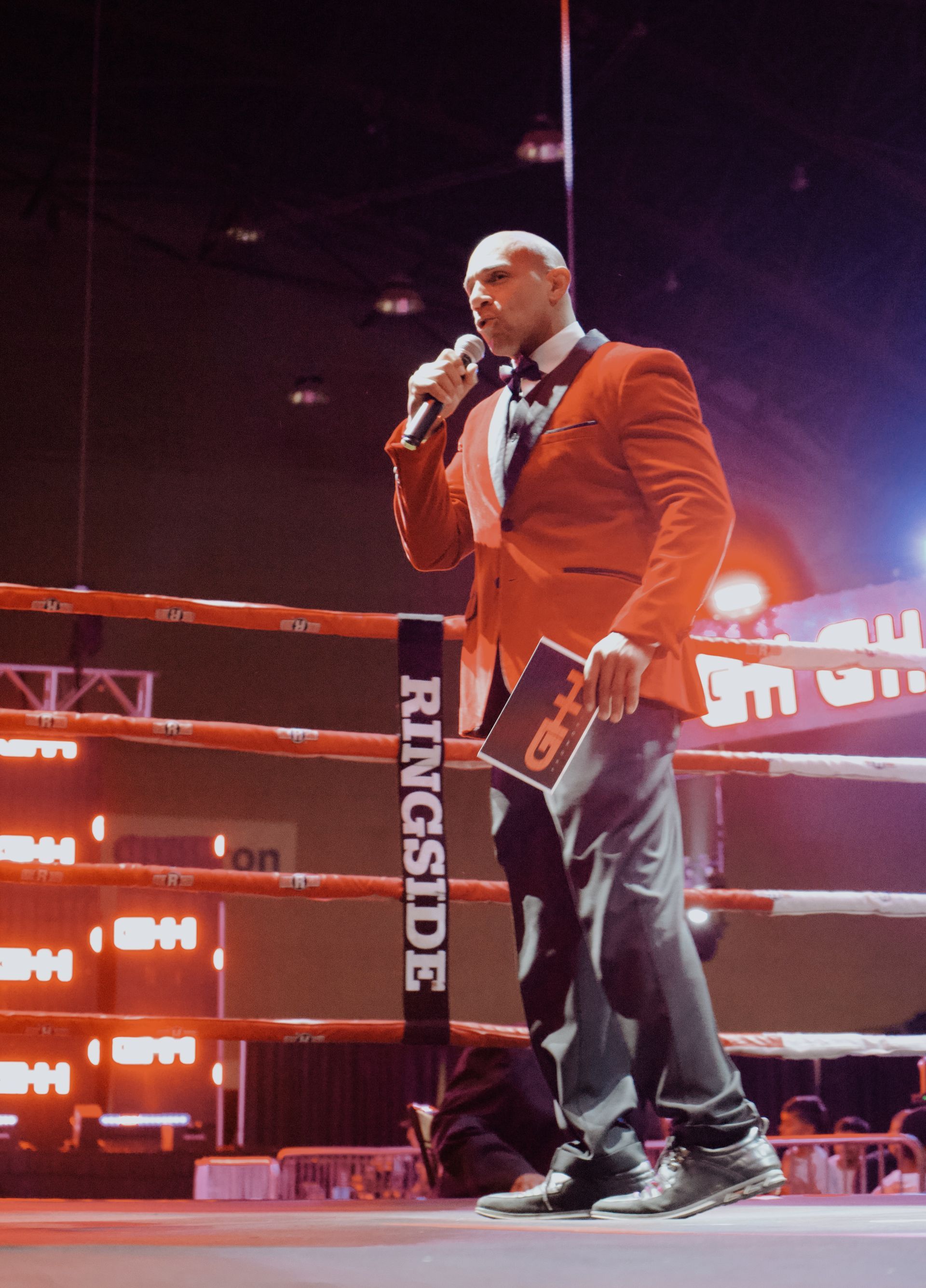 Man in a red blazer speaks into a microphone in a boxing ring at an event
