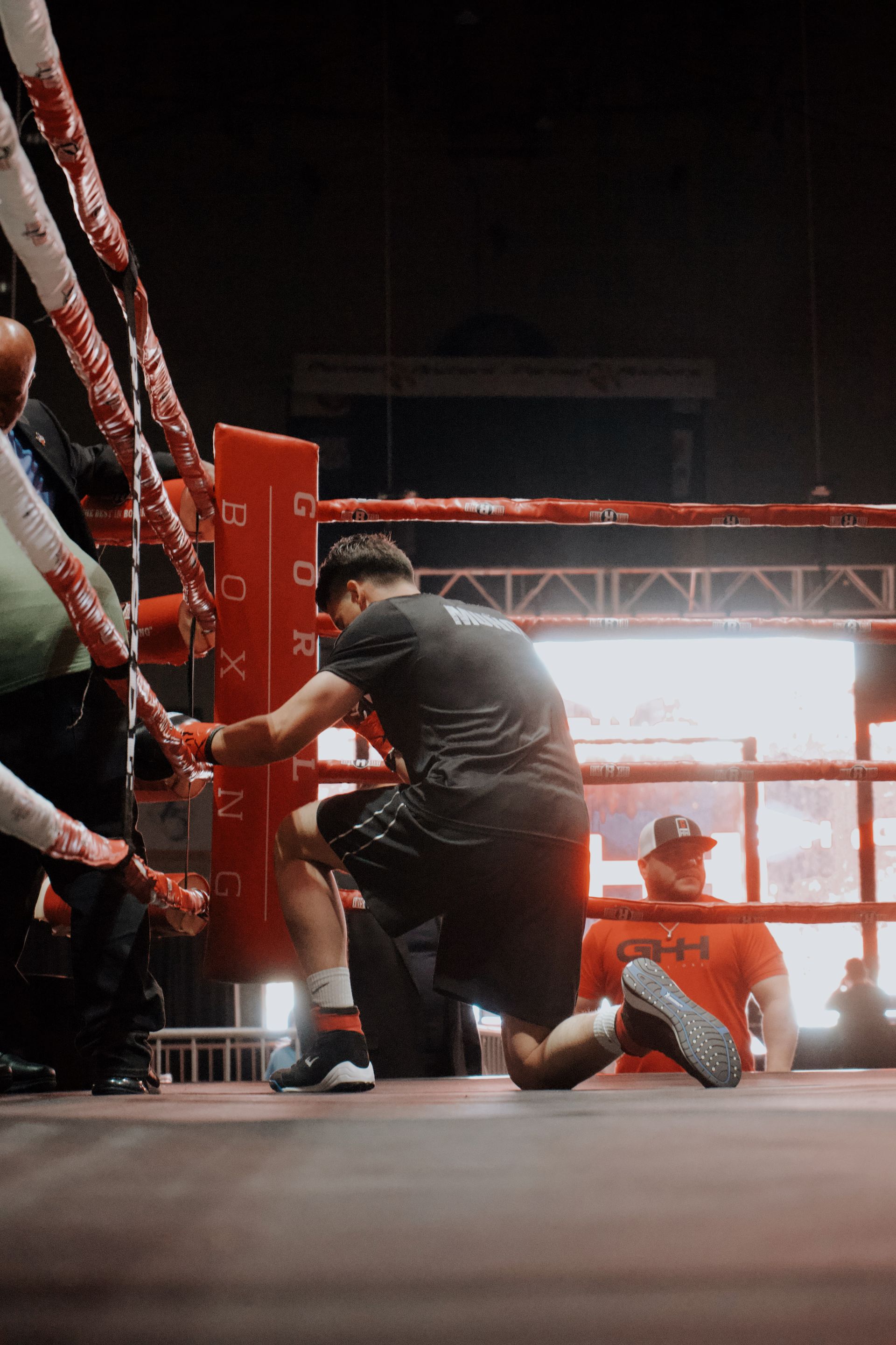 Boxers resting in a red-lit ring, one kneeling by the ropes after a match.
