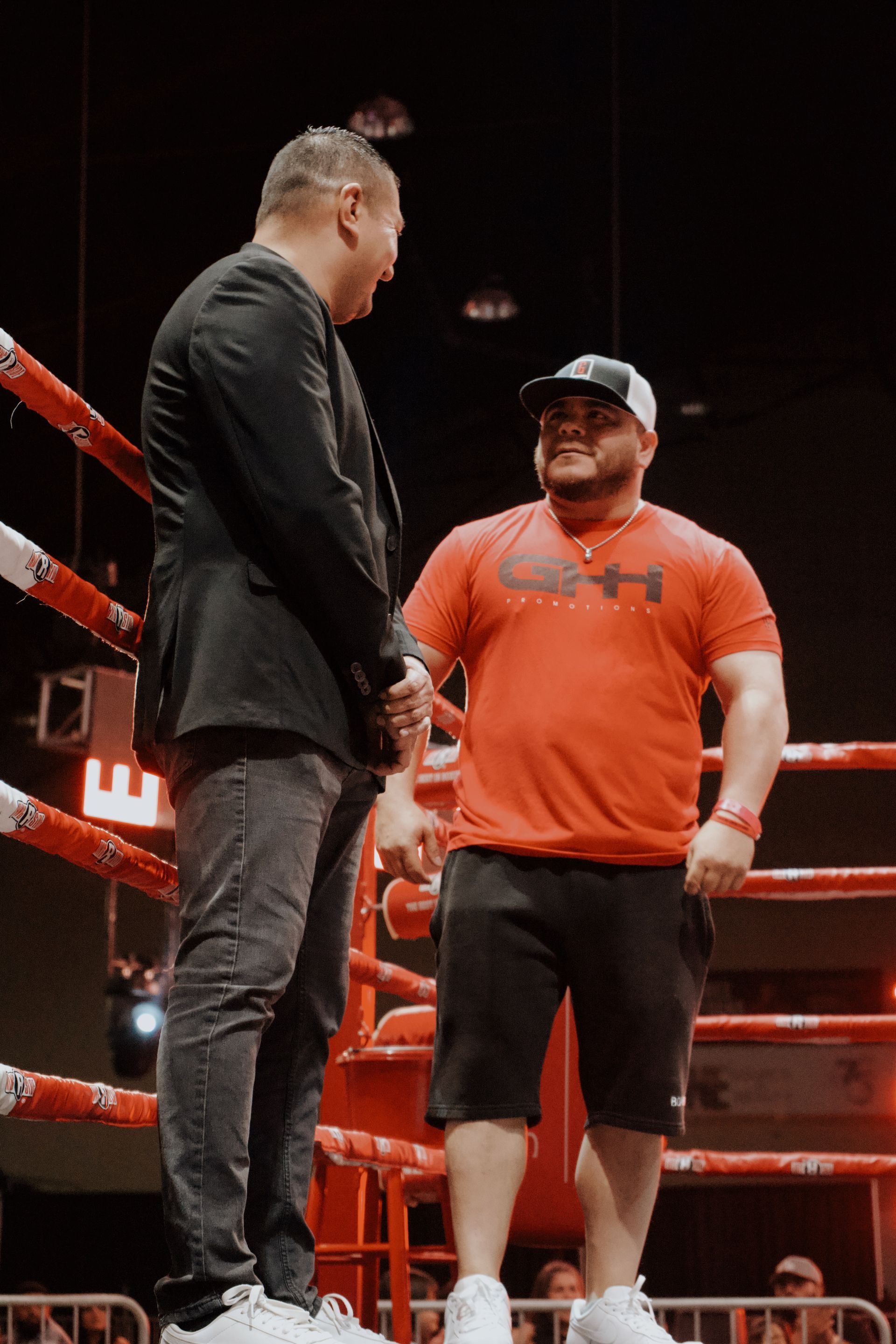 Two men face each other in a wrestling ring under red lights, one in black and one in orange.