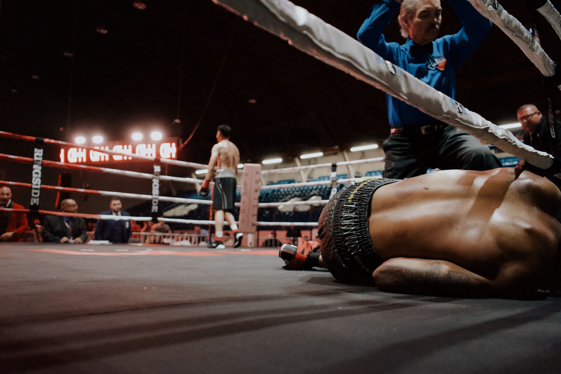 Boxing ring with a fallen fighter on the mat as the referee checks on them under bright arena lights