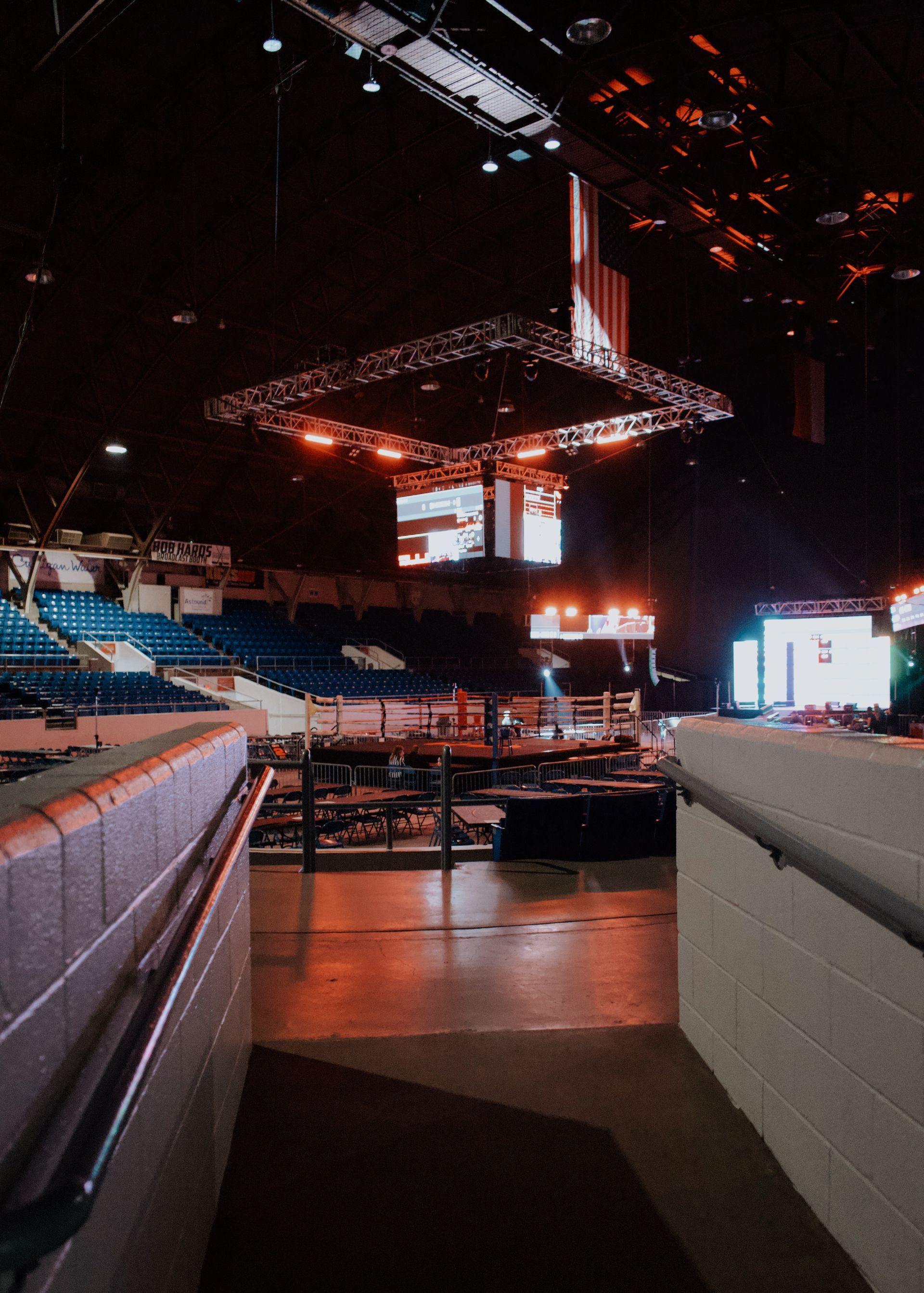 Empty indoor arena with stage lights, seating, and a large suspended screen.