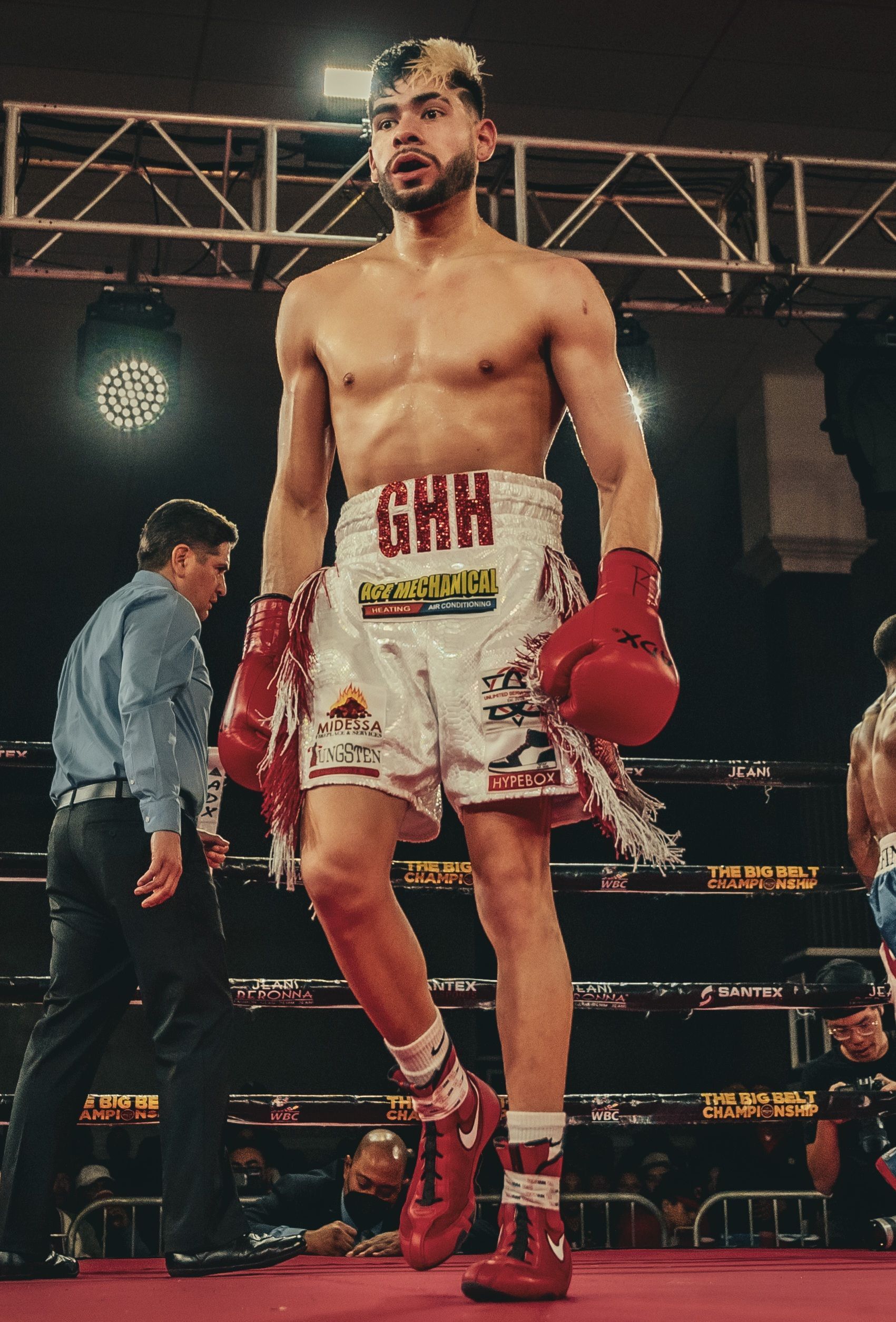 Boxer in red gloves and white trunks stands in a boxing ring under bright lights.