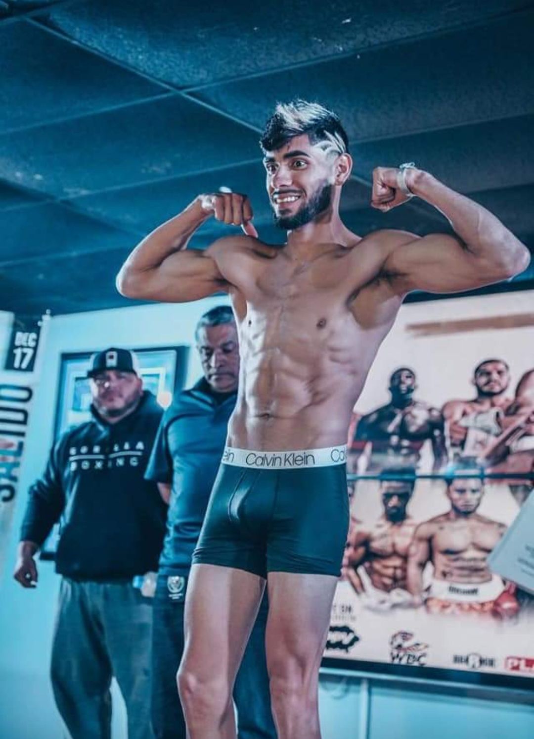 Male boxer flexing in a gym ring, wearing black trunks, with posters and a coach in the background