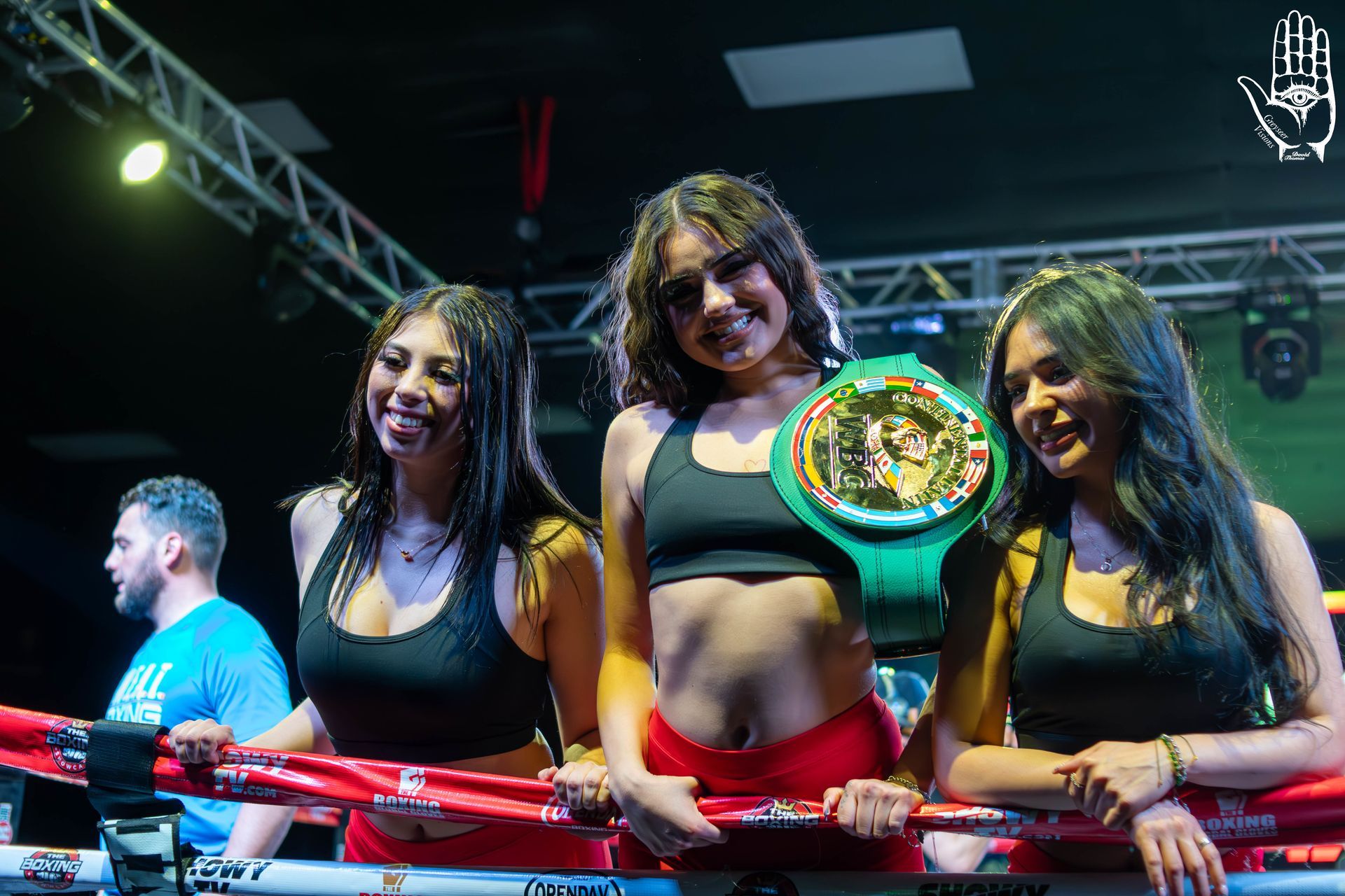 Three women at a boxing ring, smiling with a green championship belt draped over the center woman’s shoulder.
