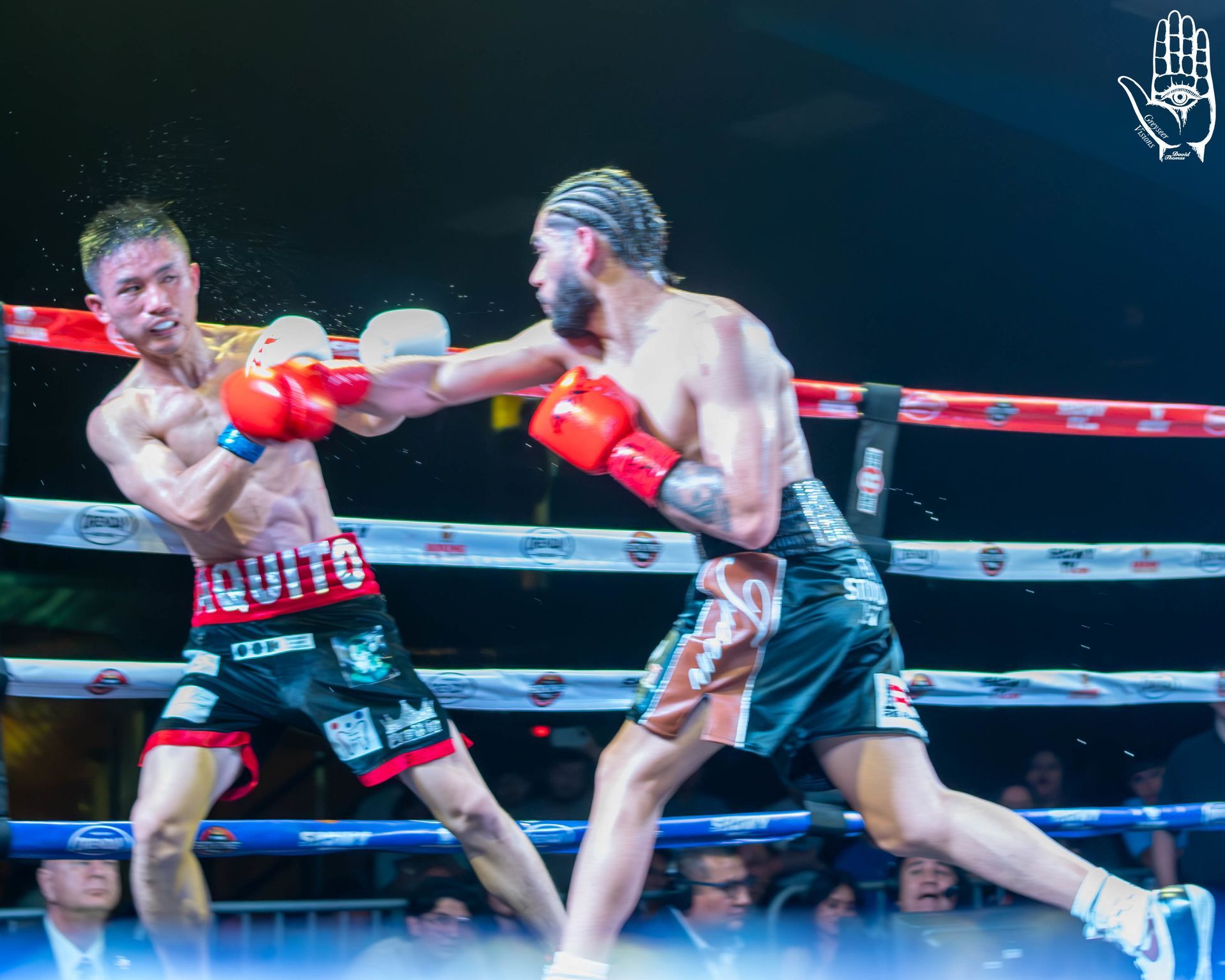 Two boxers in red gloves exchange punches in a ring under bright lights.