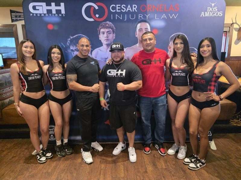 Group posing at a gym event backdrop with sponsor banners, two men centered and six women in black outfits on either side