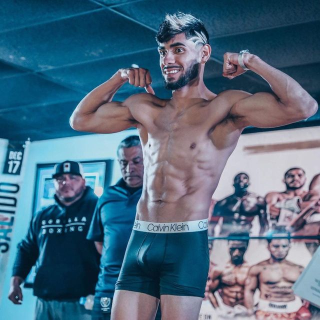Male boxer posing with flexed arms in a boxing ring, wearing black trunks and smiling under blue gym lights.