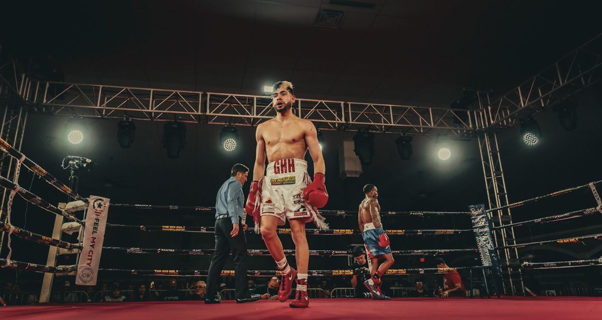 Boxers in a ring under bright lights, one in white trunks centered in front of a red canvas