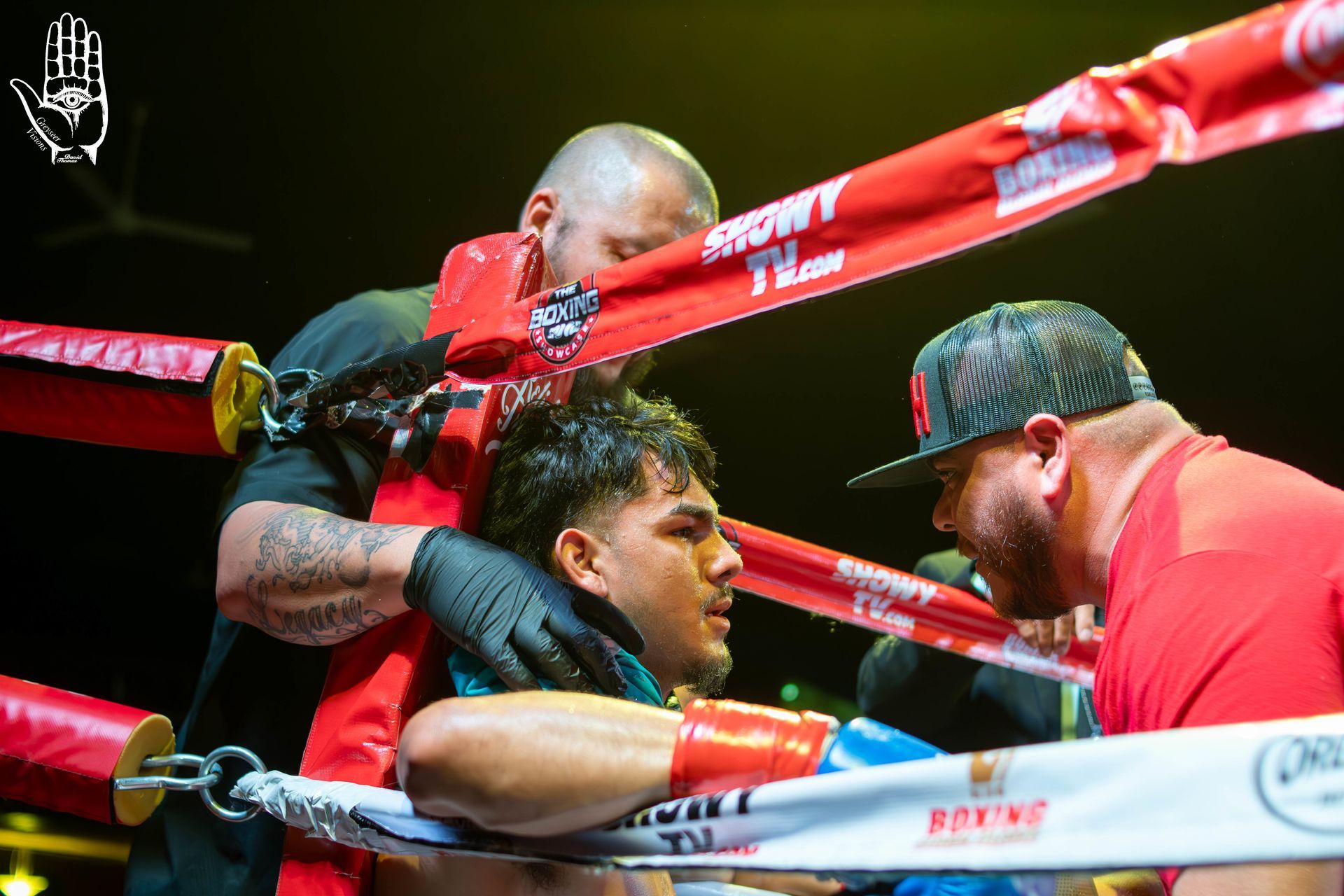 Boxers in a ring between red and white ropes during a match, with coaches leaning in from the corners