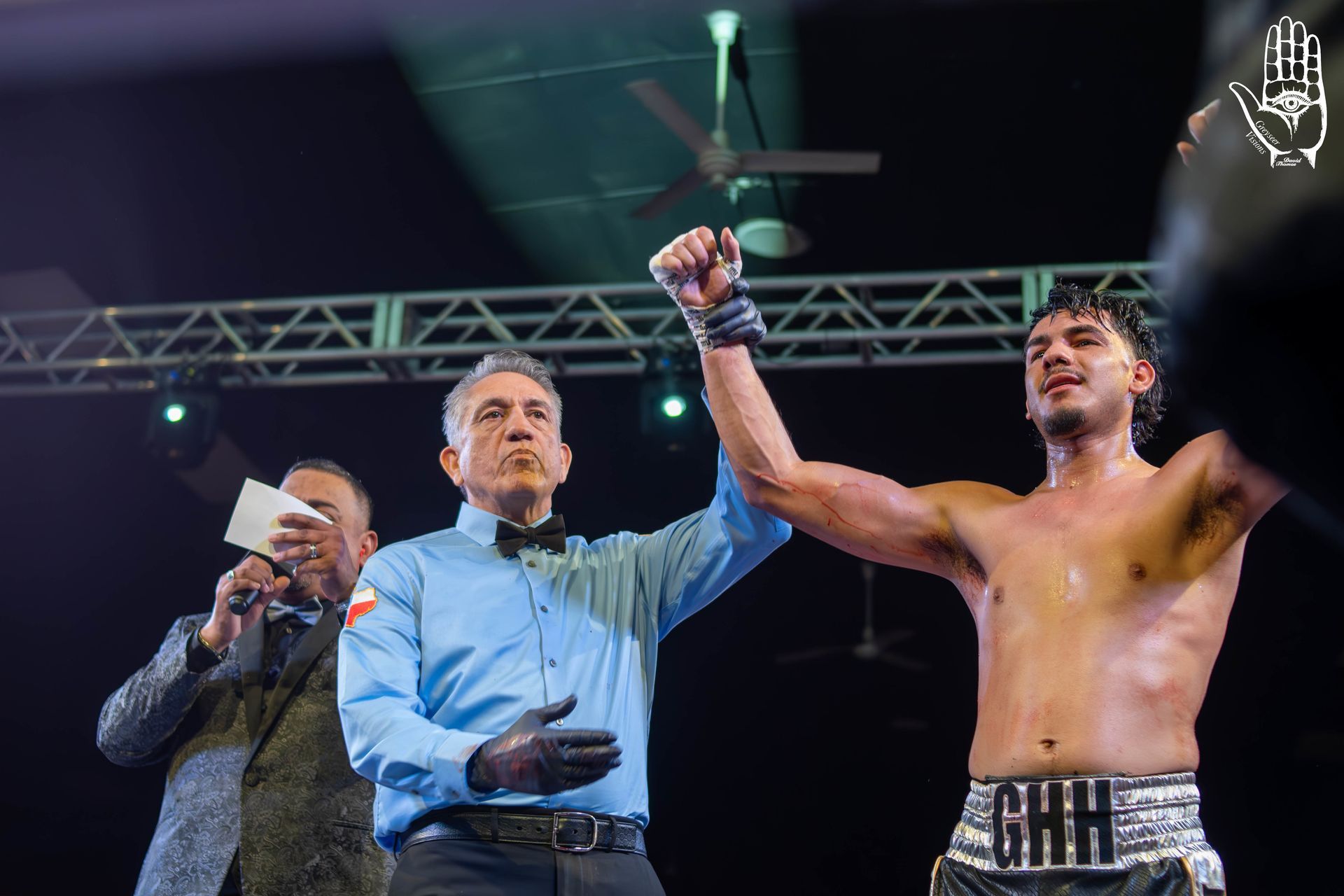 Boxing referee raises a fighter’s hand in victory under arena lights, with announcer nearby.