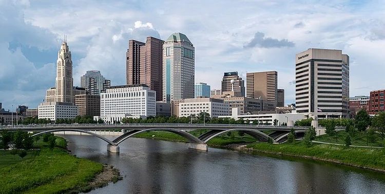 City skyline with tall buildings beside a river and arched bridge under a partly cloudy sky.