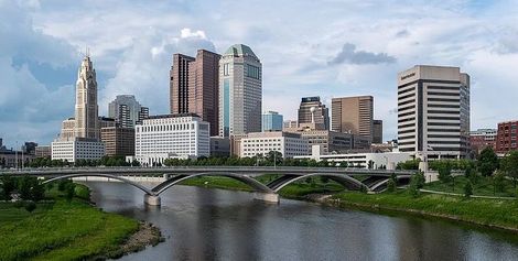 City skyline with tall buildings beside a river and arched bridge under a partly cloudy sky.
