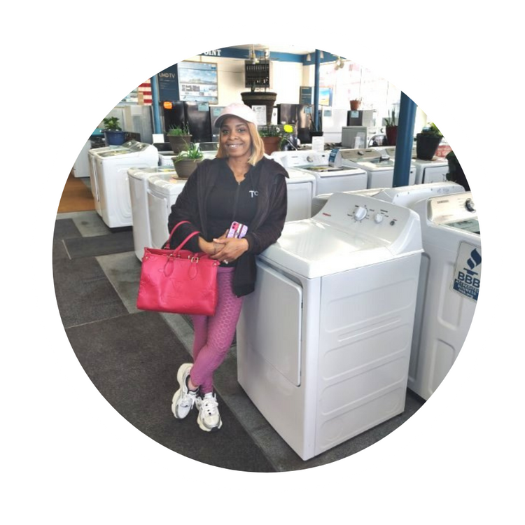 Woman leaning on a washing machine in an appliance store, holding a bright pink handbag.