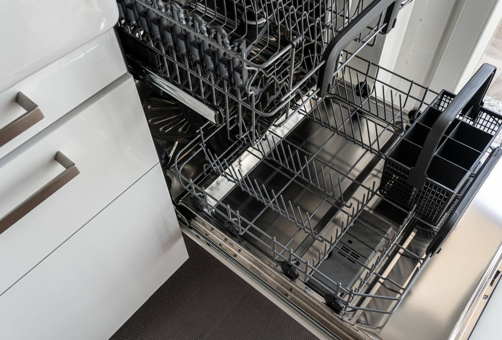 An open dishwasher with empty racks, showcasing a metal interior and a black cutlery basket.