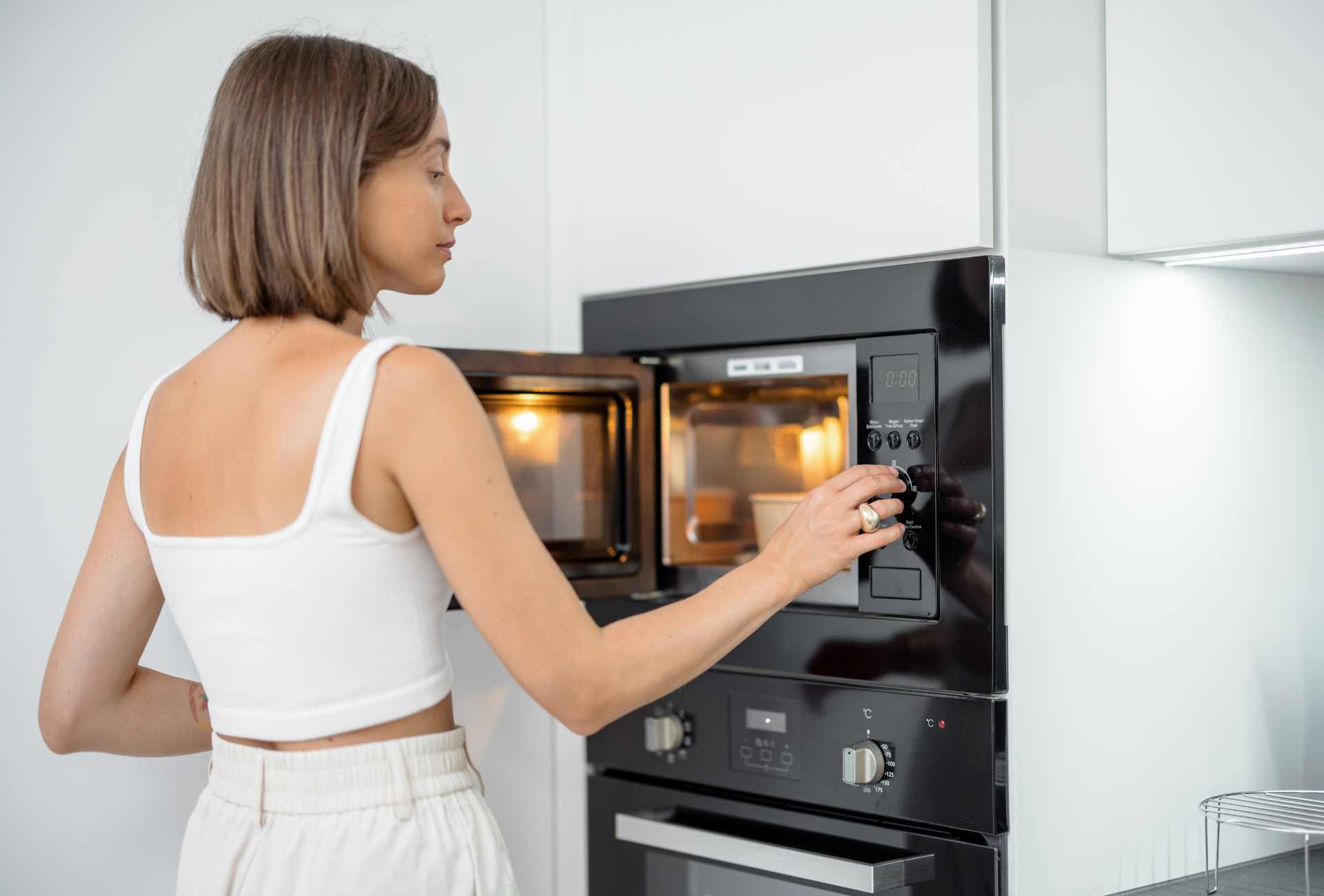 Woman opening a microwave in a white kitchen, preparing food.