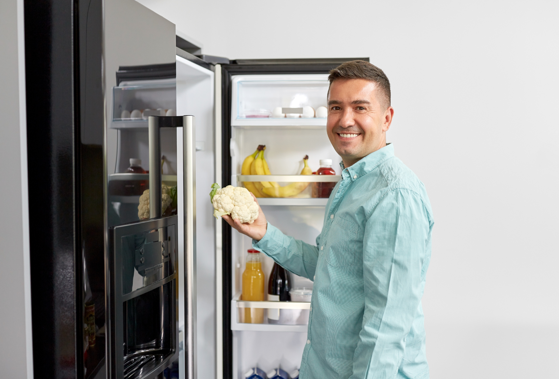Man holding cauliflower in front of open refrigerator, smiling.