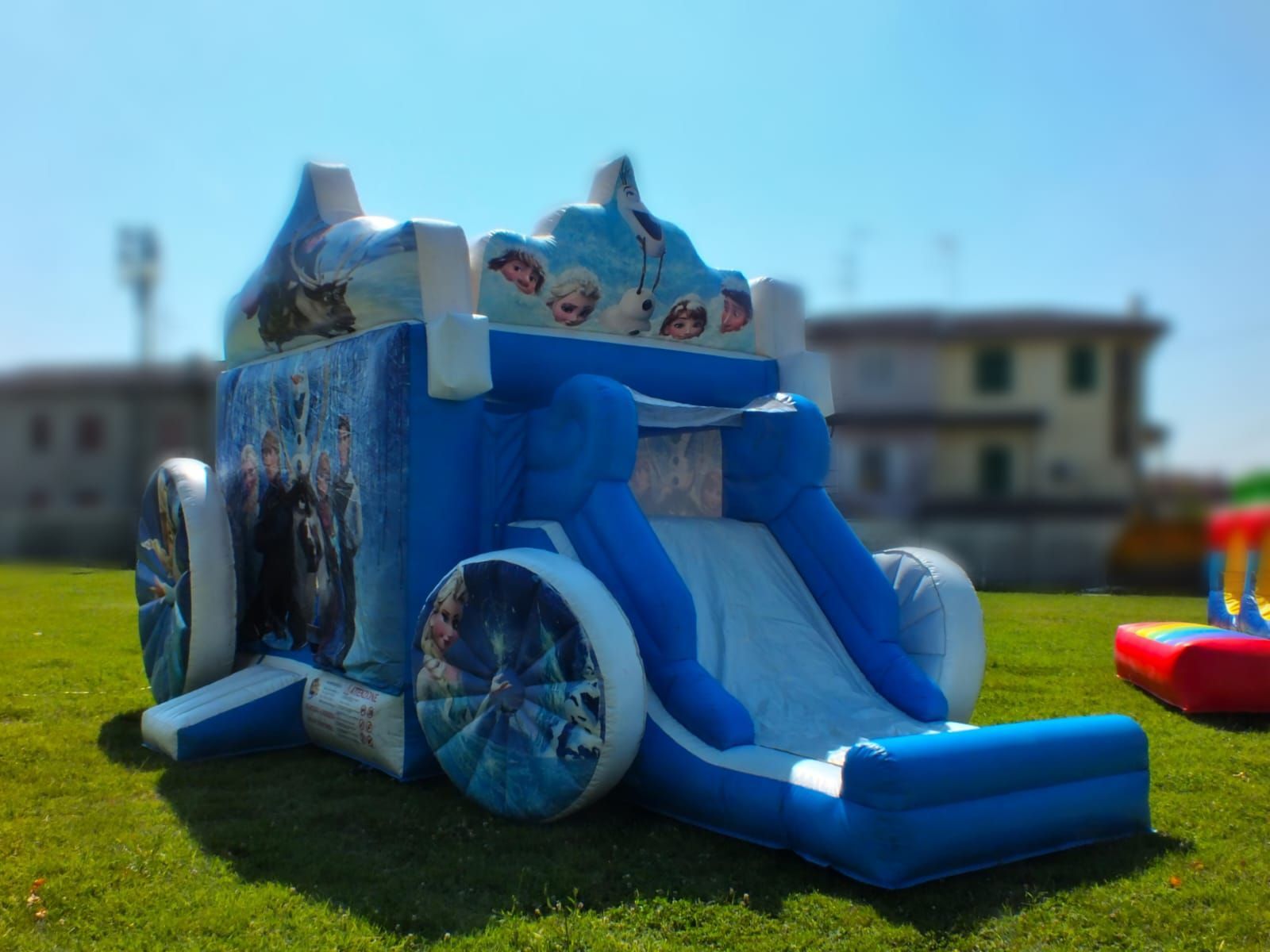 Castello gonfiabile blu con scivolo su un prato, con un edificio sfocato sullo sfondo.