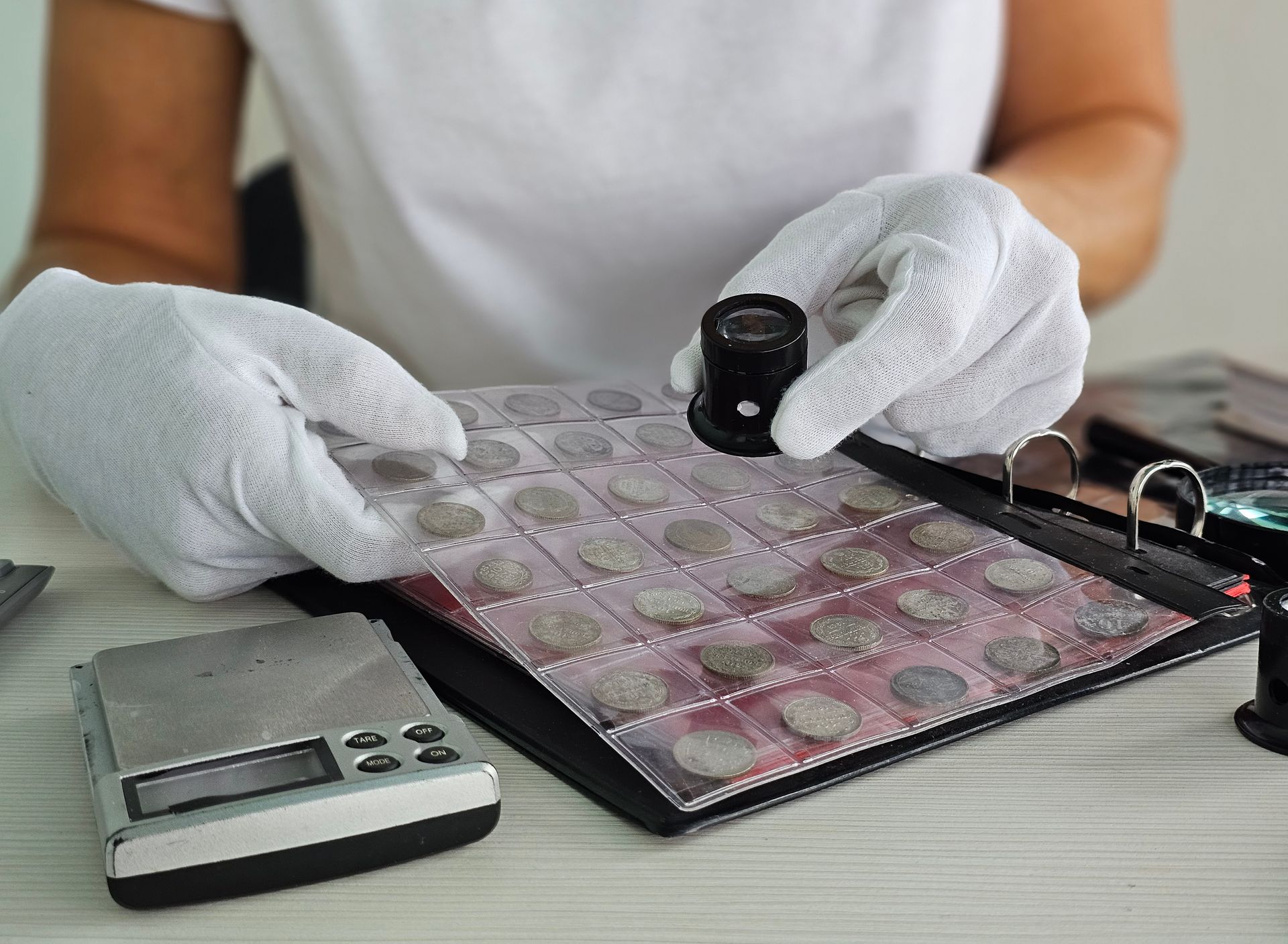 Gloved collector examines coins with a loupe near a digital scale.