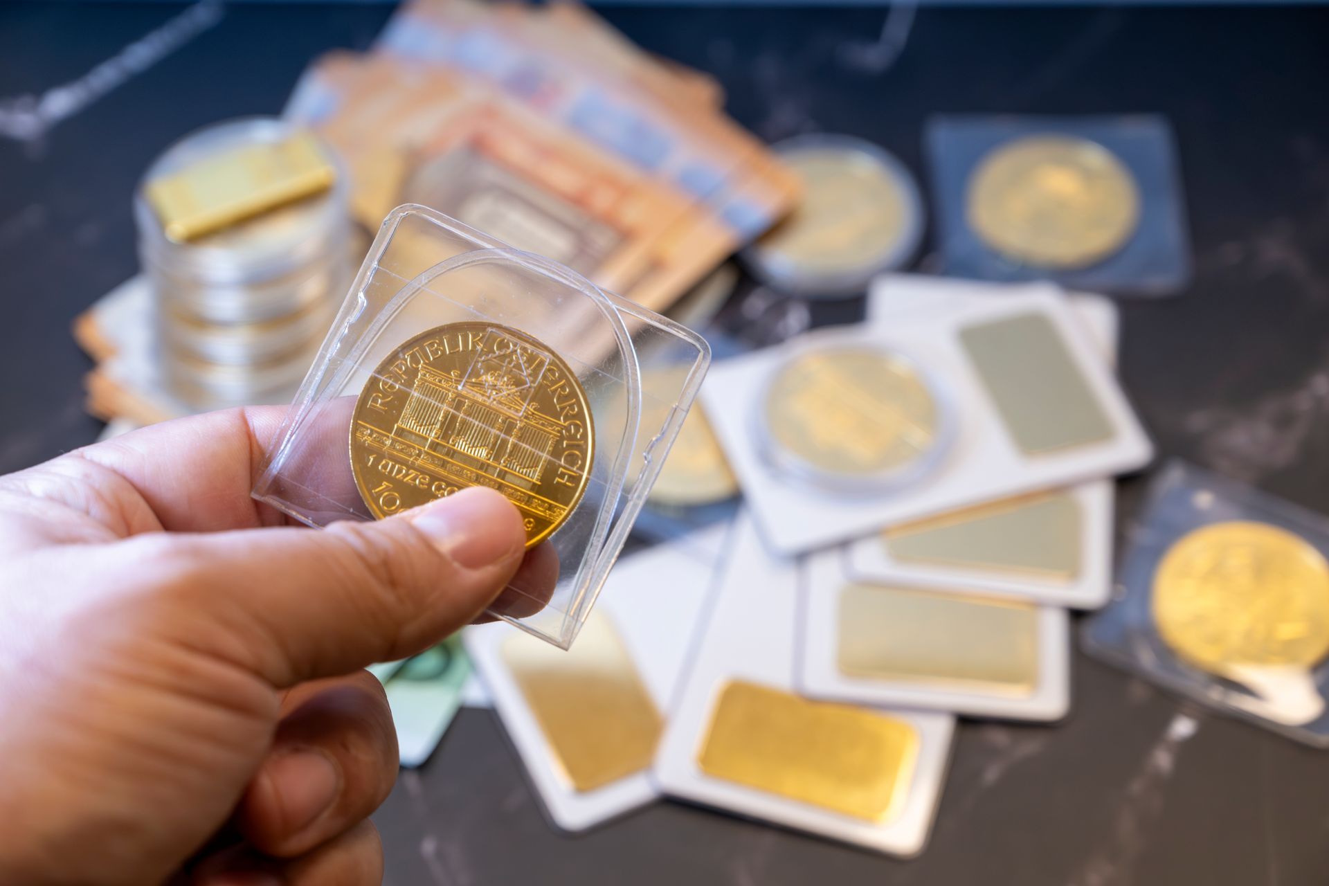 Hand holding a gold coin in a case with more coins and bars on a table. Hand holding a gold coin in a case with more coins and bars on a table.