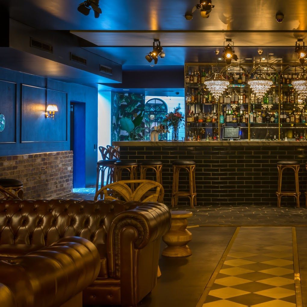 Dimly lit lounge with brown leather seating, a tiled floor, and a bar lined with glasses and warm lights.