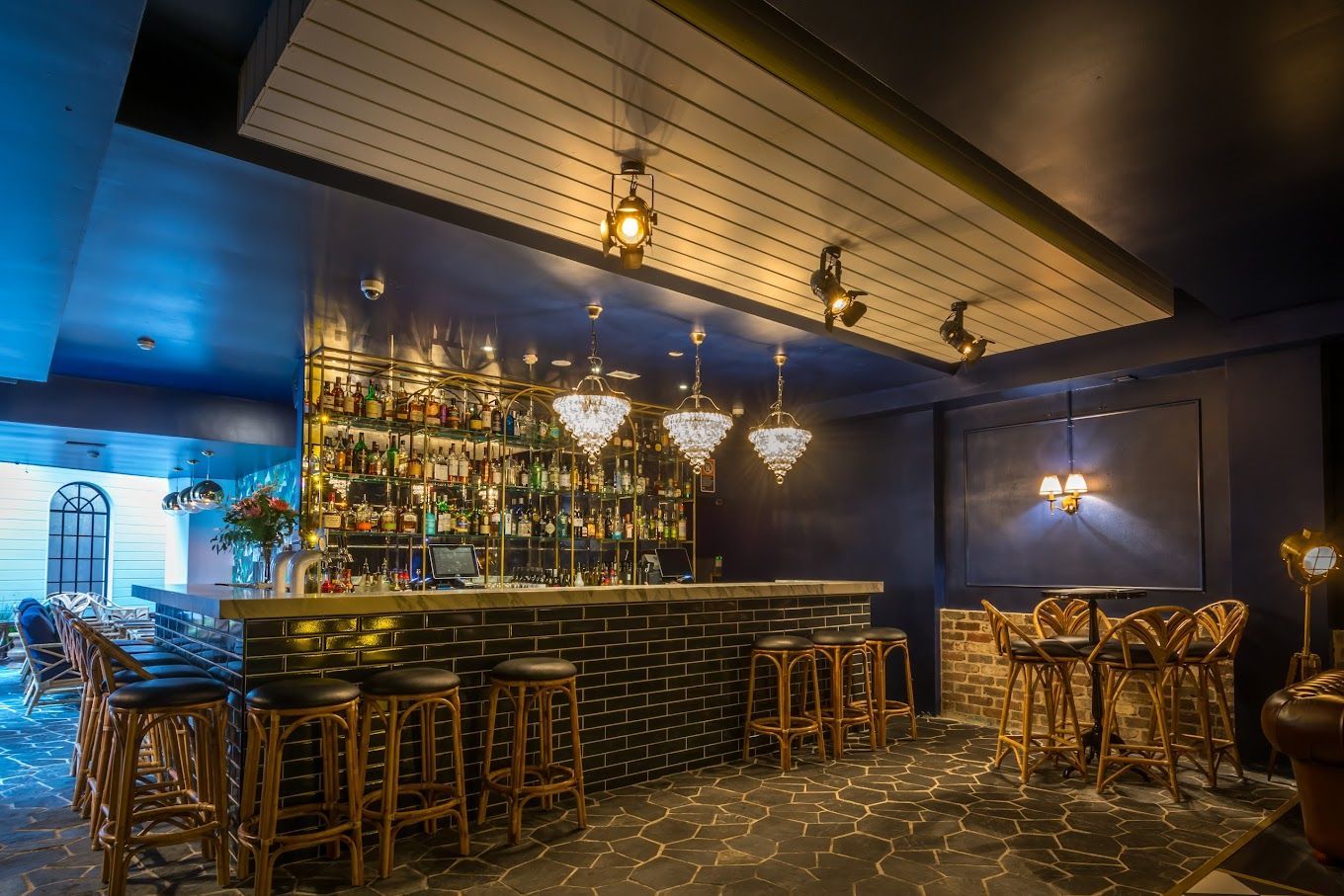 Dimly lit modern bar interior with gold accents, hanging lights, shelves of bottles, and bar stools