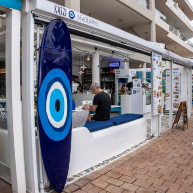 Beachside kiosk with blue eye sign, white counter, and a man working inside.