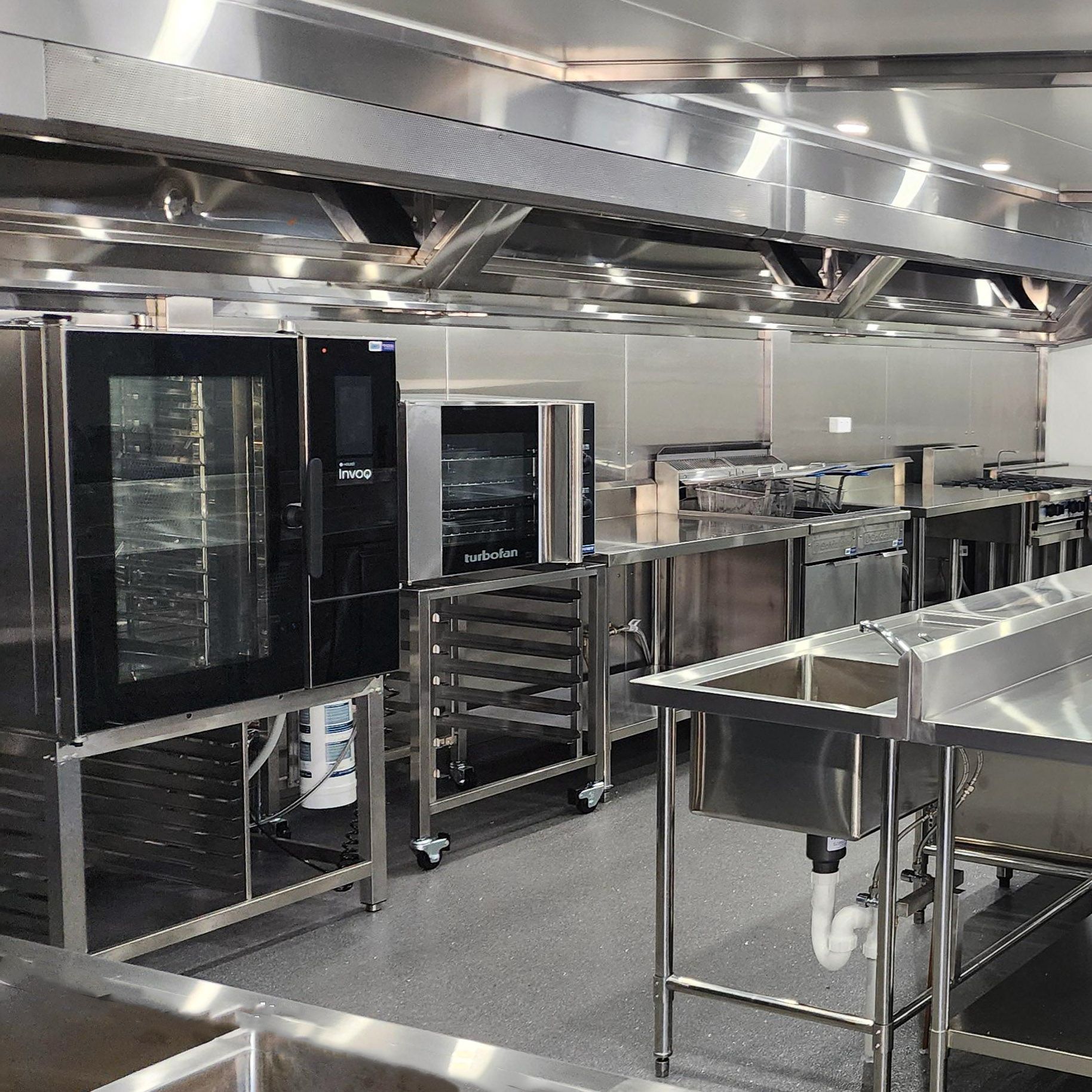 Stainless-steel commercial kitchen with ovens, racks, and prep tables under bright lights
