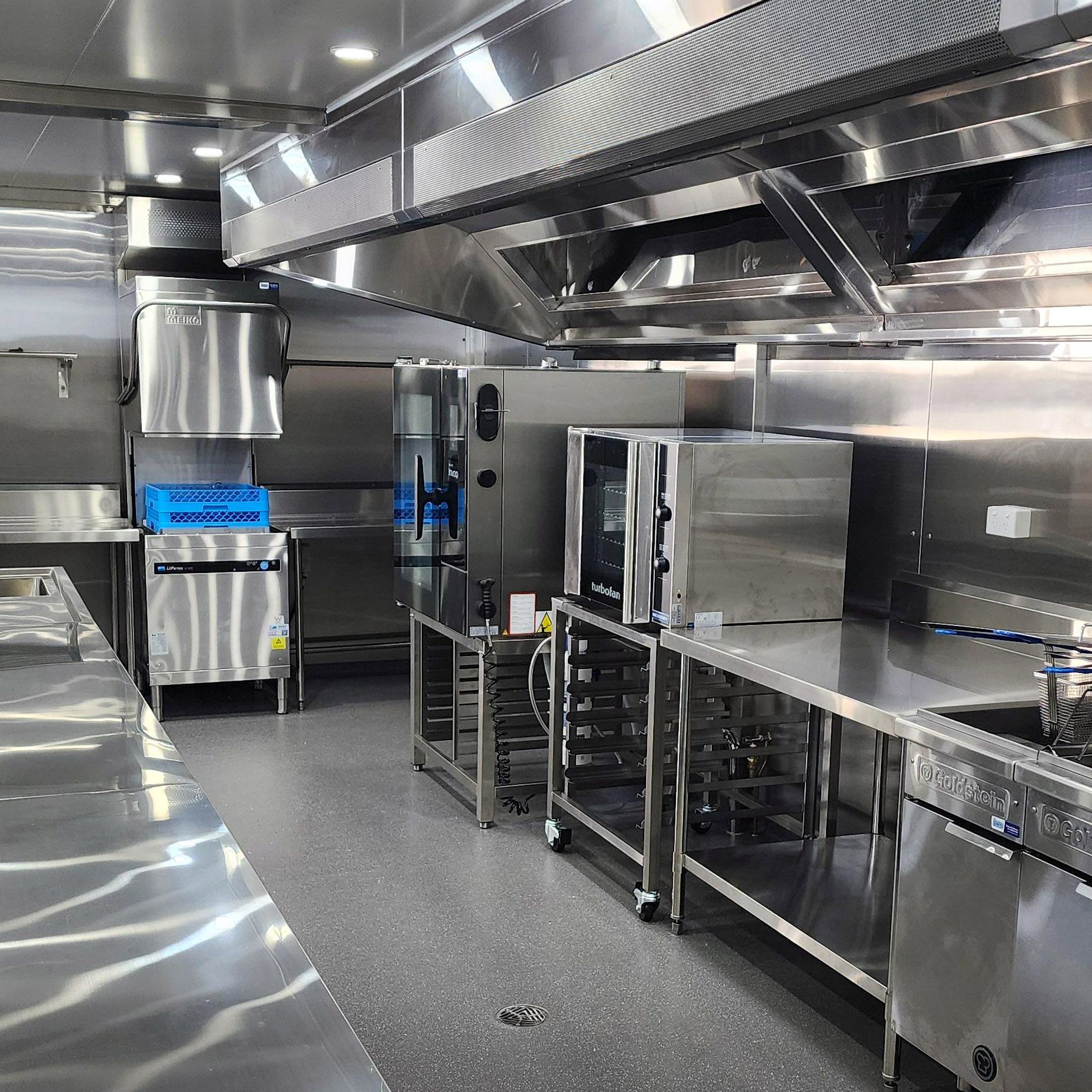 Stainless steel commercial kitchen with counters, ovens, and overhead ventilation.