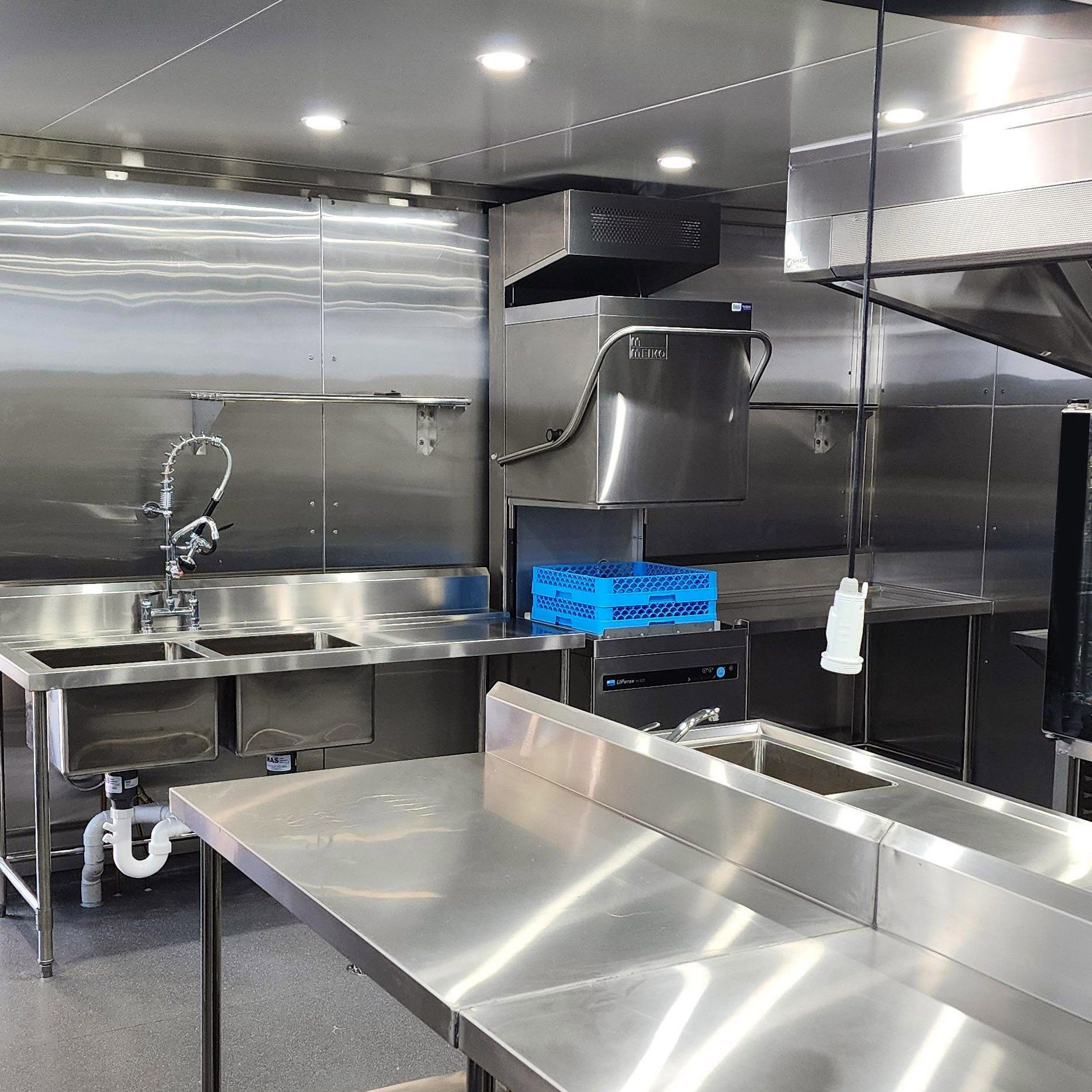 Stainless steel commercial kitchen with sinks, counters, range hood, and bright overhead lights