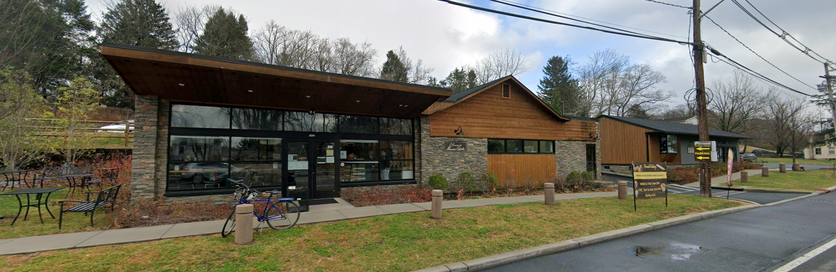 Modern glass-and-wood building beside a wet road under overcast skies, with trees and utility lines nearby