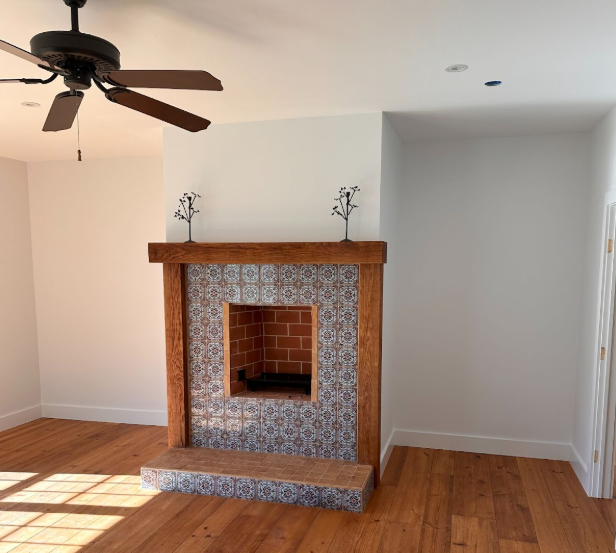 Empty living room with brick fireplace, hardwood floors, and a ceiling fan in daylight.