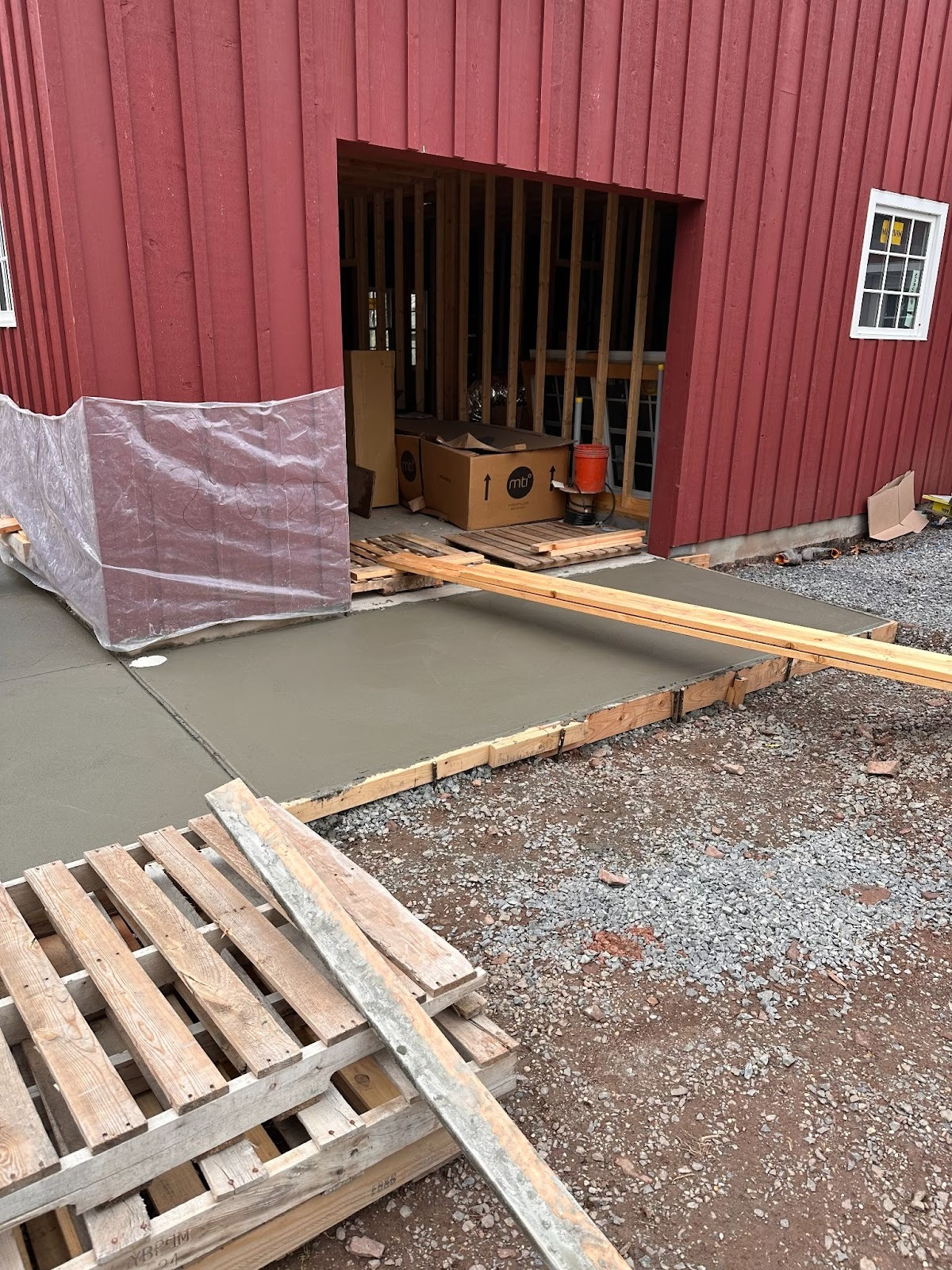 Fresh concrete ramp leading to a red barn doorway, with pallets and gravel in front.