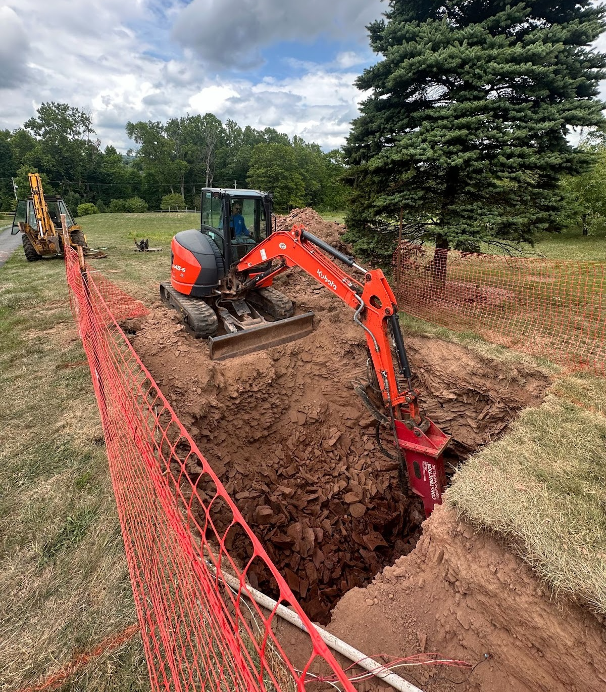 Orange excavator digging a deep trench beside a red safety fence in a grassy field