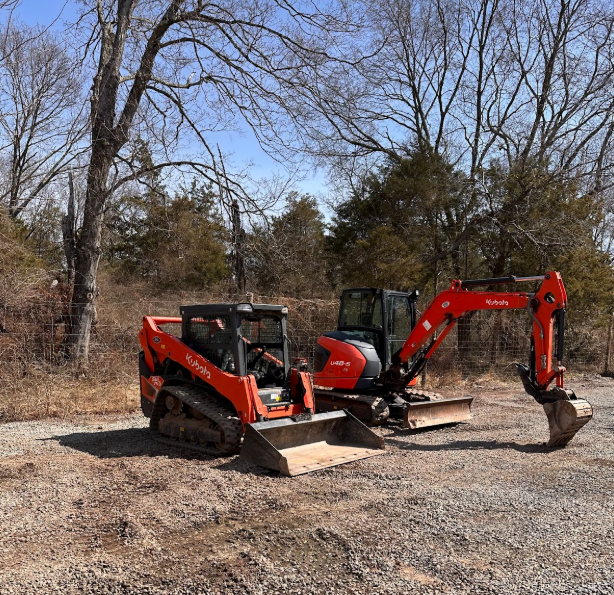 Two red excavators parked on a dirt lot near leafless trees under a blue sky