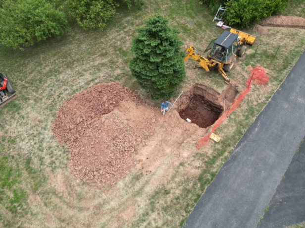 Aerial view of backhoe digging a large hole beside a driveway, with a pile of dirt and a tree nearby