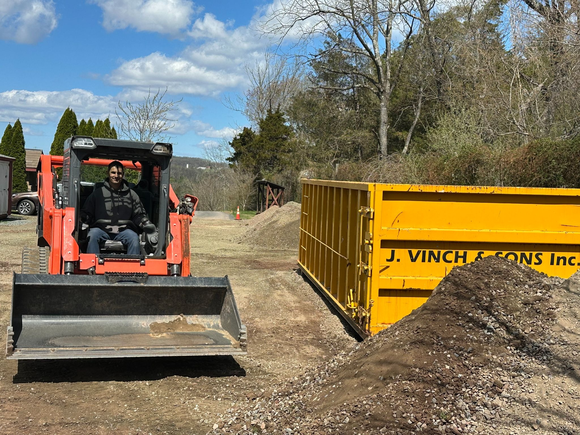 Orange skid steer on dirt road beside a yellow dumpster and brush pile under a blue sky