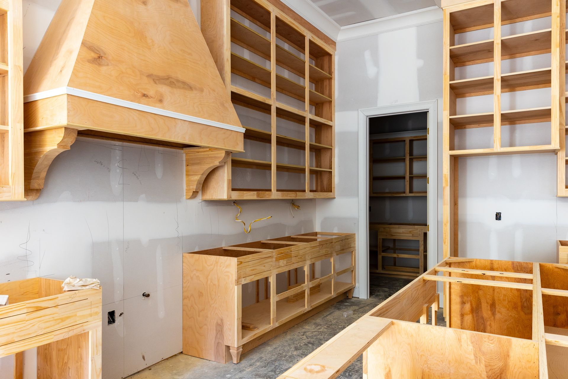 Unfinished kitchen with wooden cabinets and shelves installed, white walls, and a large range hood.