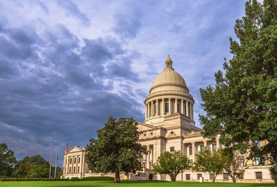 Dome of a neoclassical capitol building under a dramatic cloudy sky, framed by trees and lawn
