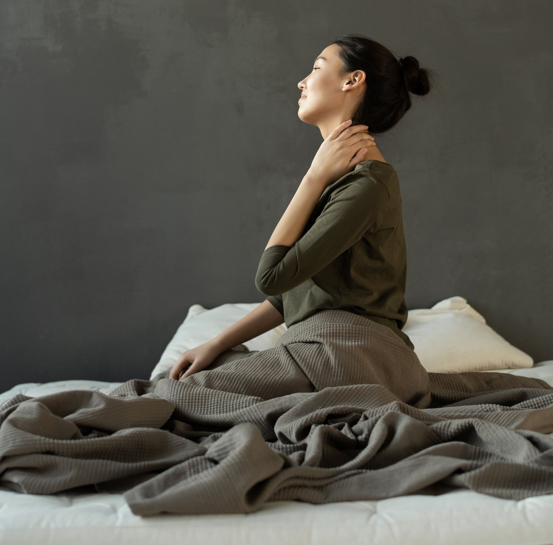 a woman sits on a bed with her eyes closed and her hand on her neck