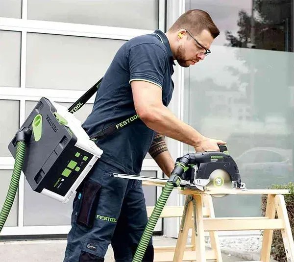 Worker using a circular saw to cut wood at a workbench, with a green power tool backpack nearby