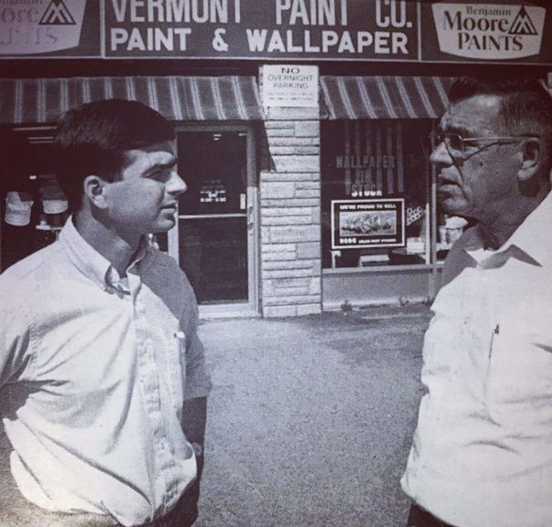 Two men talking outside Vermont Paint & Wallpaper store, with storefront signs in the background.