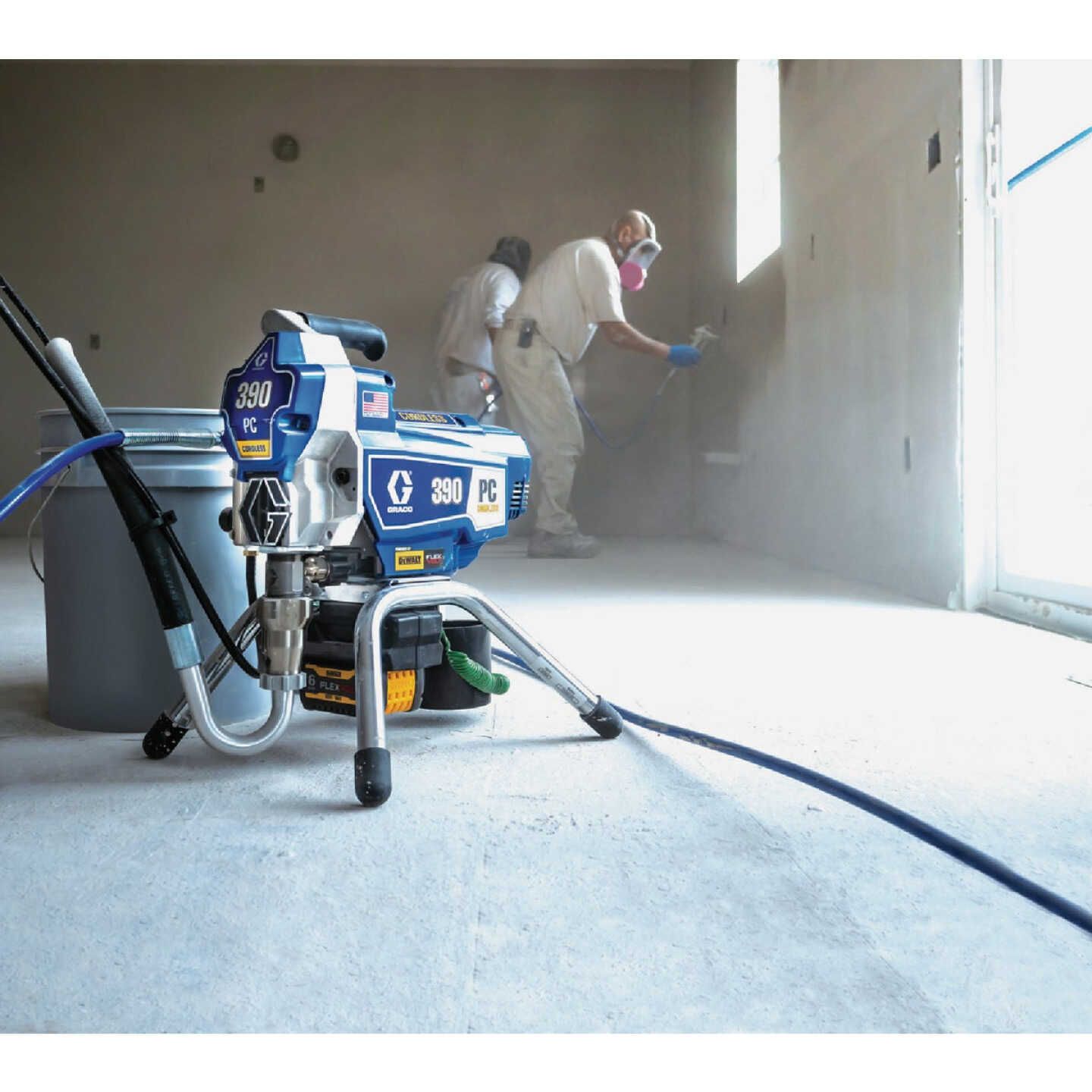 Workers spray painting a wall inside a bright room, with a blue paint sprayer in the foreground.