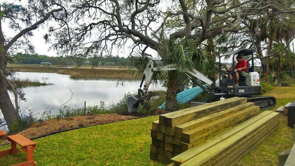 Excavator beside a pond with stacked lumber and a person working under a large tree